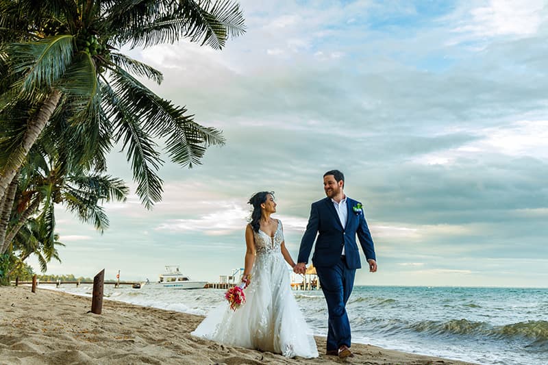 A newlywed couple walks hand in hand along a sandy beach, surrounded by palm trees under a cloudy sky. A newlywed couple walks hand in hand along a sandy beach, surrounded by palm trees under a cloudy sky.