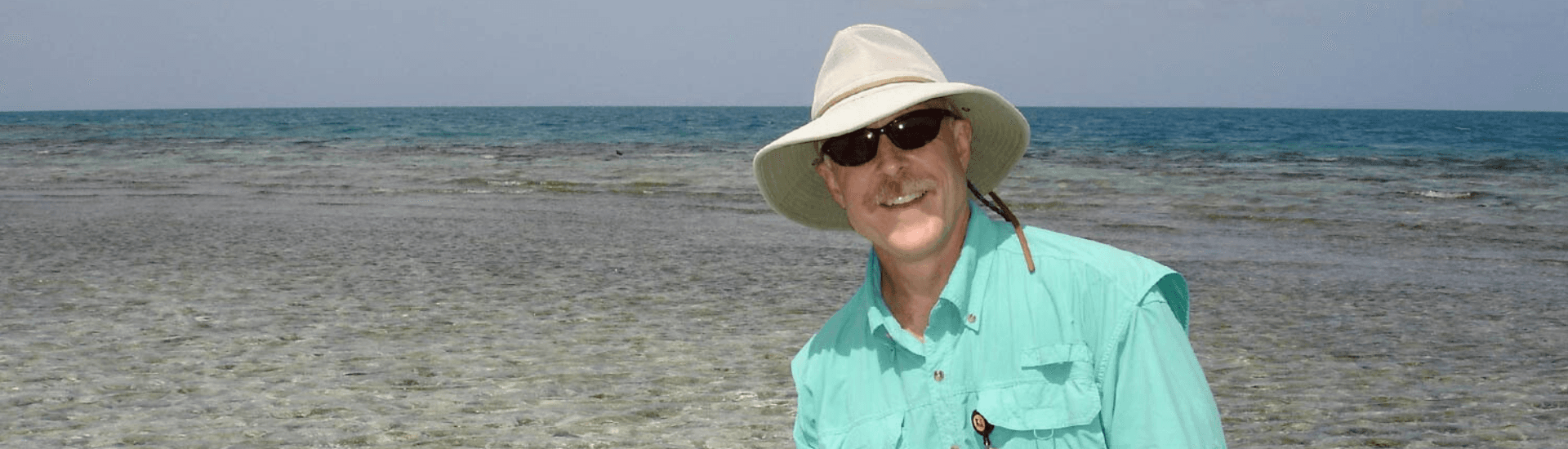 A man in a fishing hat and green fishing shirt smiles widely while backed by the beach and ocean.