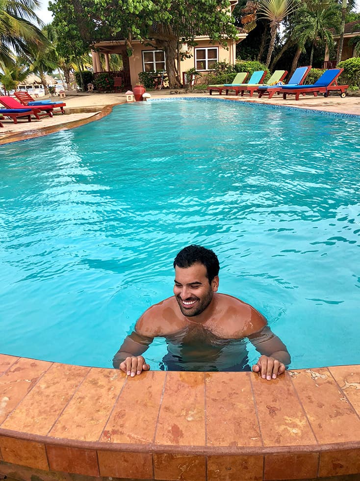 Dark haired man standing in pool with pool chairs surrounding the deck.