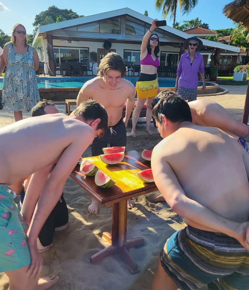 A group of young people crowded around a table in the sand, focused on a watermelon-eating contest, while others watch from a nearby pool area.