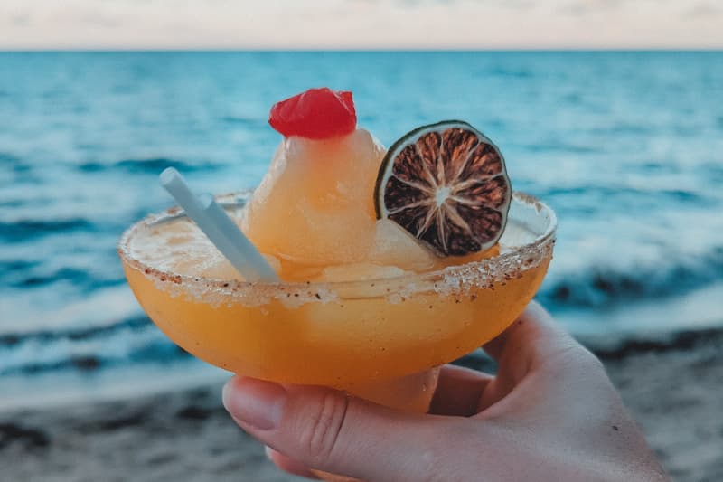 A hand holds a tropical cocktail with a garnish, against a beach and ocean backdrop. A hand holds a tropical cocktail with a garnish, against a beach and ocean backdrop.