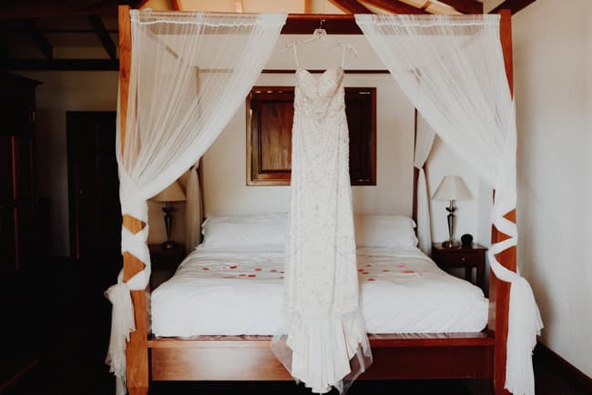 A wedding dress hangs from a wooden canopy bed in a softly lit bedroom.