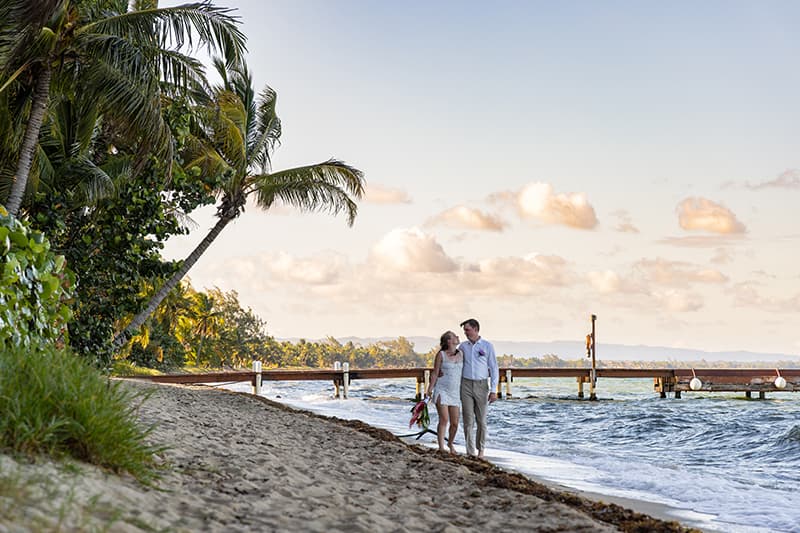 A couple walks hand-in-hand along a sandy beach, with palm trees and a pier in the background.