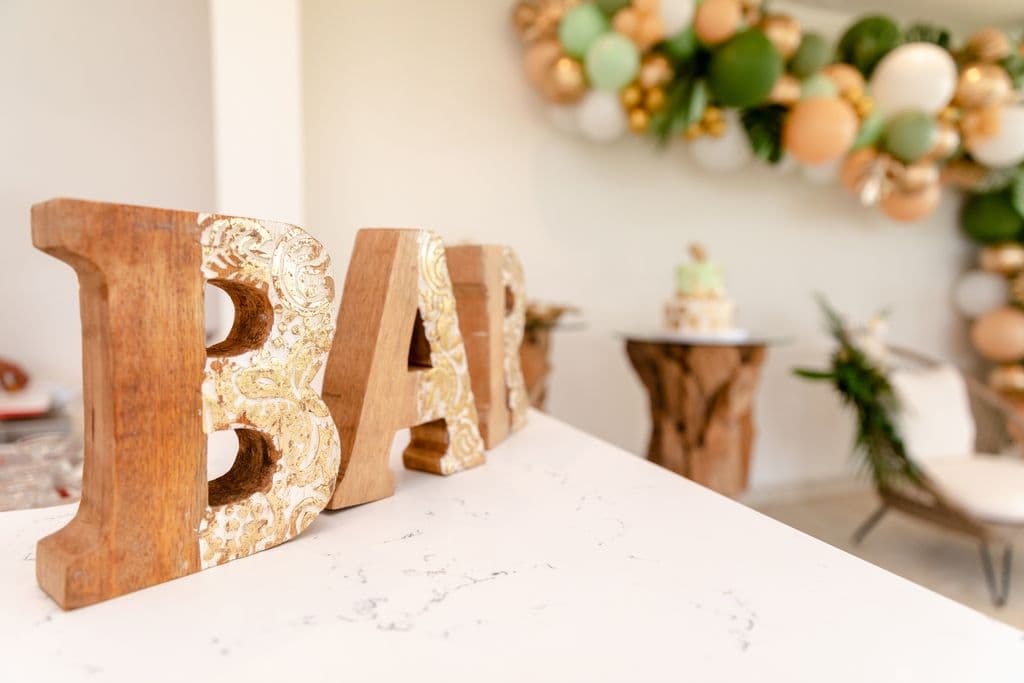 Wooden letters spelling "BAR" with gold accents, displayed on a table at a baby shower. A balloon garland and a cake are visible in the background.