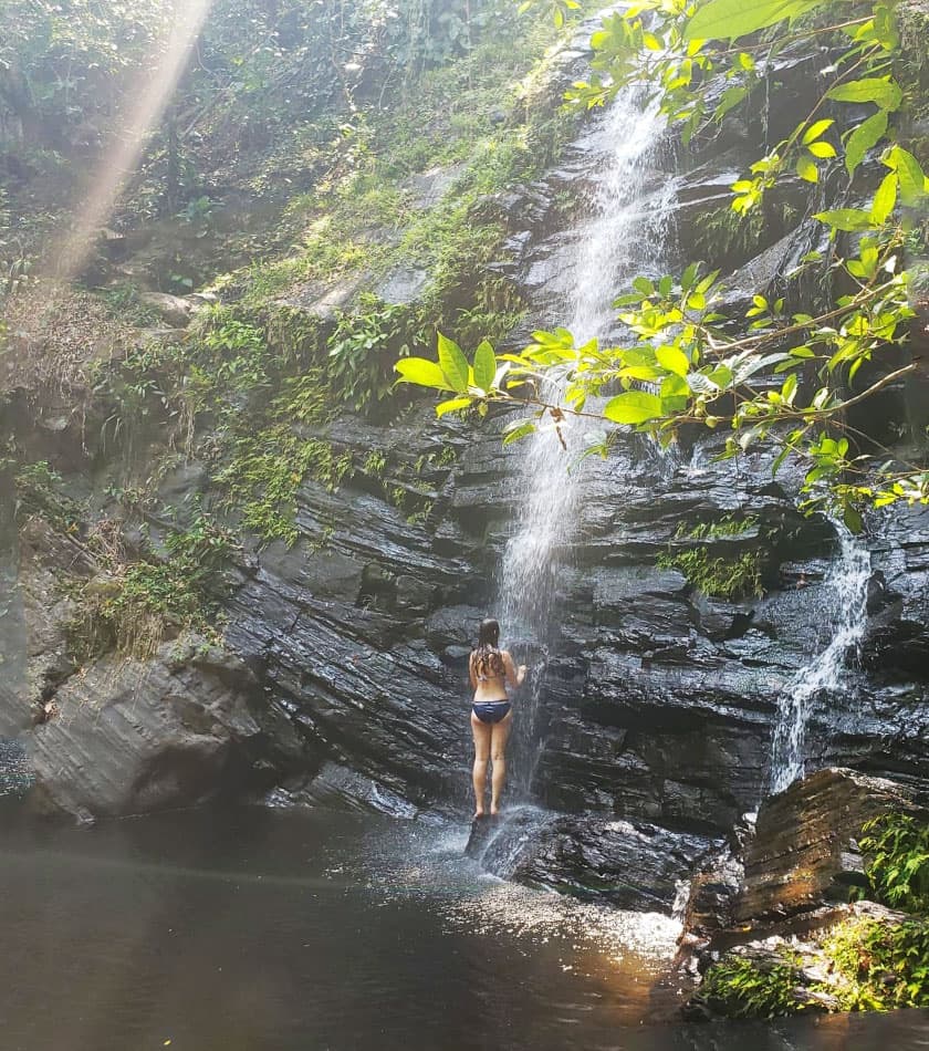 A person stands under a waterfall, surrounded by lush greenery and rocky terrain.