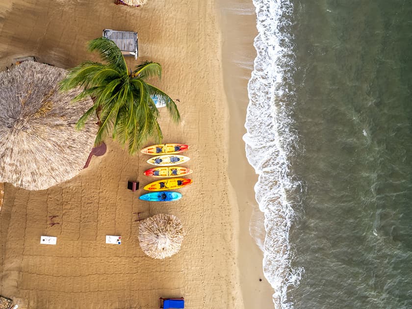 Aerial view of a sandy beach with colorful kayaks, palm trees, and straw huts near the water's edge.