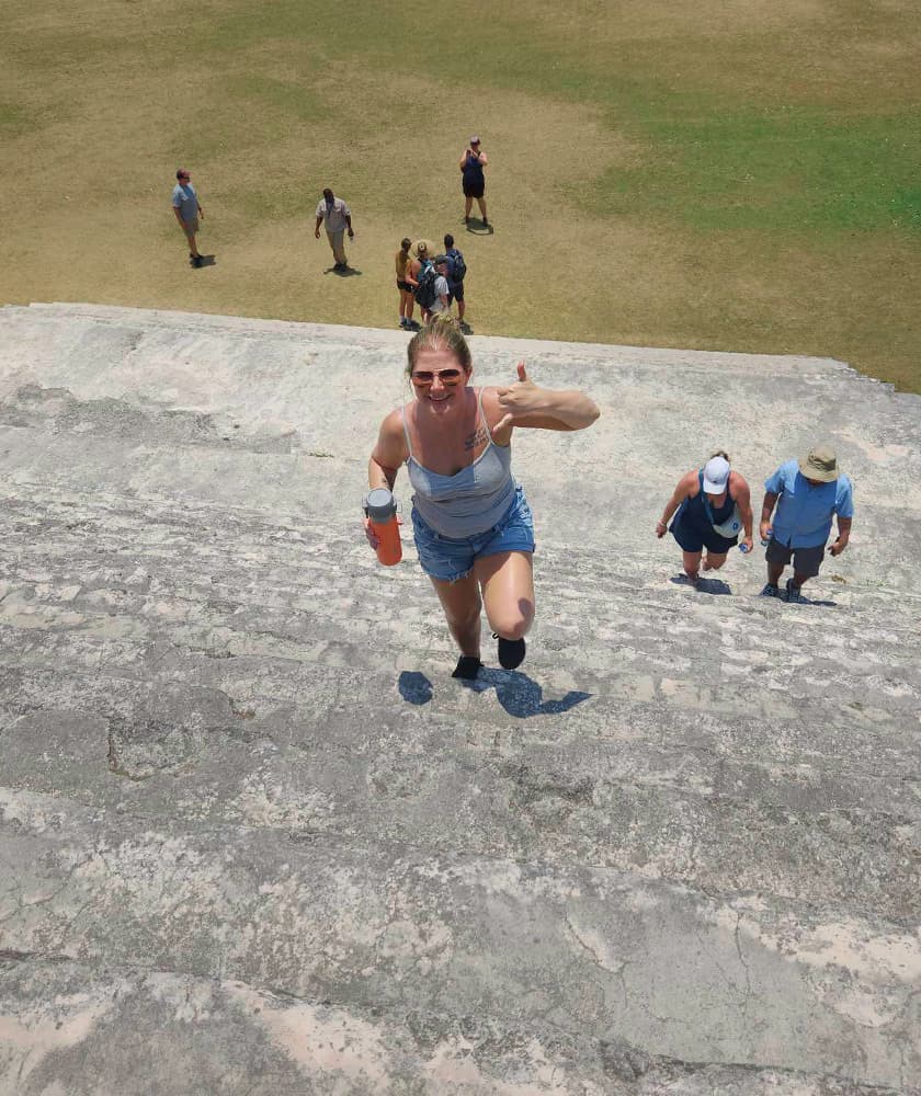 A woman climbs stairs at a historic site, smiling and holding a drink, with others in the background.