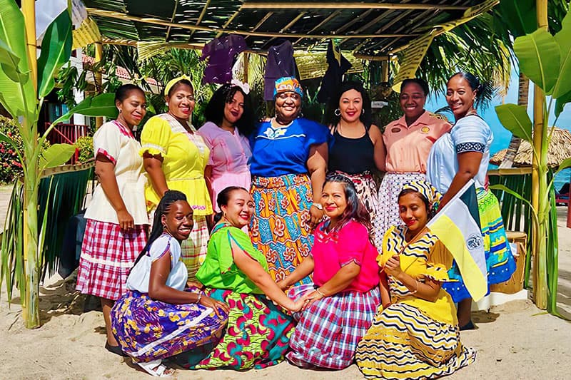 A group of women in colorful traditional attire poses together under a palm-frond structure near the beach. A group of women in colorful traditional attire poses together under a palm-frond structure near the beach.