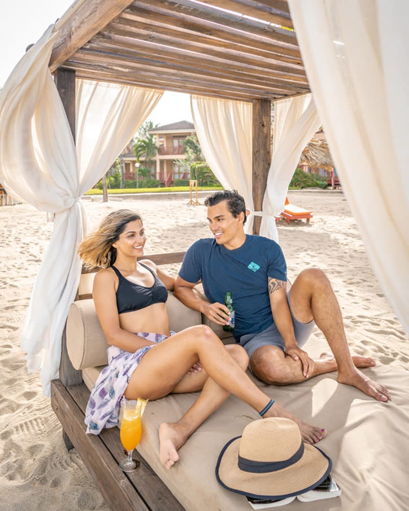 A couple enjoys drinks while lounging on a beach cabana.