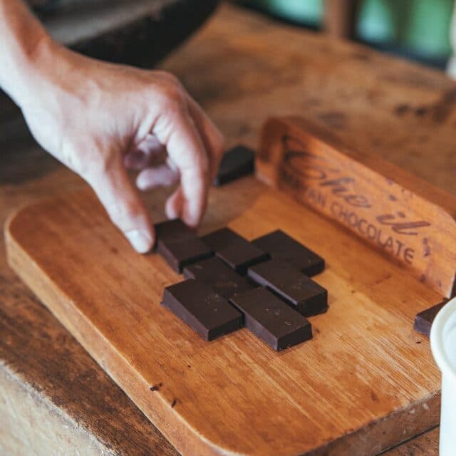 A person's hand reaches out to pick up a square of dark chocolate from a wooden cutting board. The chocolate is arranged in rows on the board, and there is a wooden sign with the words "The Hill Chocolate" in the background.