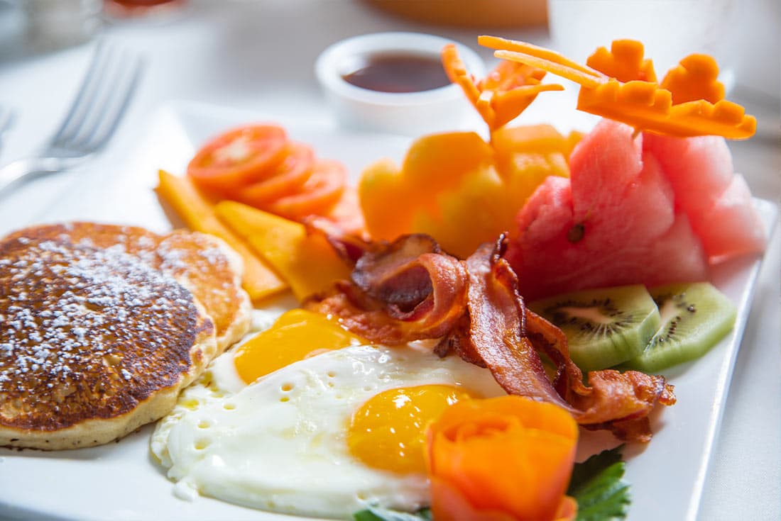 A delicious breakfast plate featuring pancakes, fried eggs, bacon, fruit, and a side of hash browns. The fruit includes watermelon, kiwi, and mango, and the eggs are perfectly cooked with runny yolks.