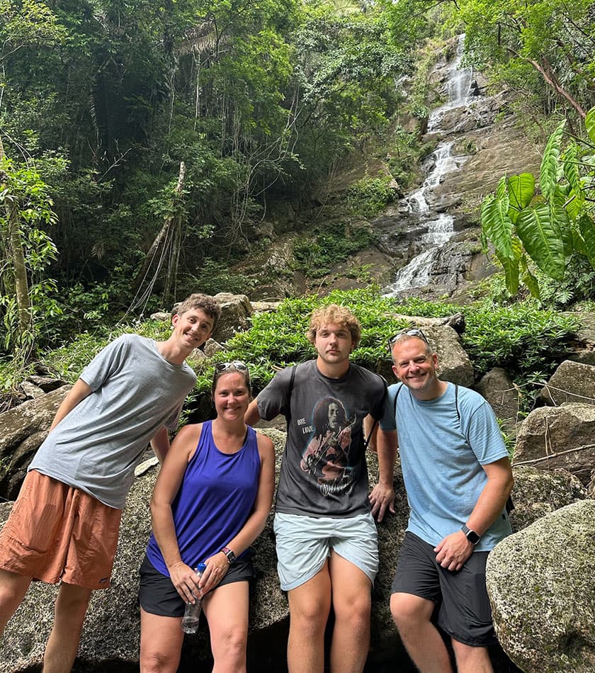 A group of four people stands on a rocky outcrop in front of a lush, green waterfall.