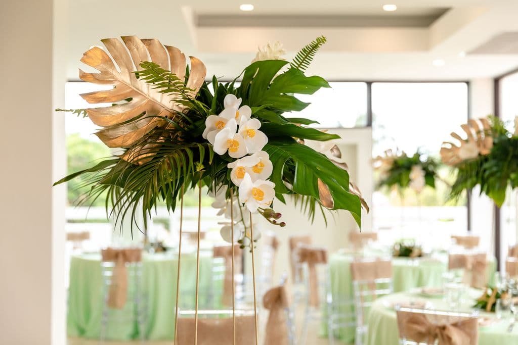 A tall floral centerpiece with white orchids, green ferns, and gold-leafed monstera leaves. The centerpiece is standing on a metal stand in a room with round tables covered in green tablecloths.