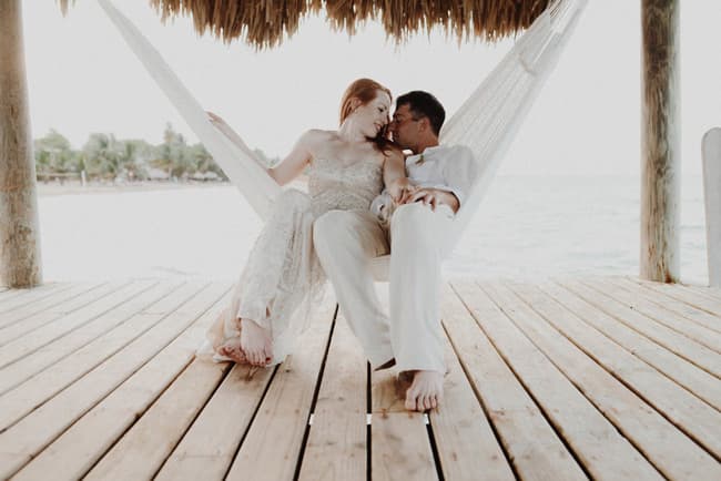 A couple in white clothing relaxes together on a hammock by the beach.
