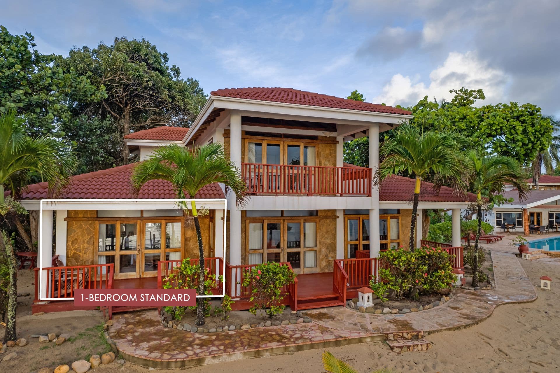 The image shows a 1-bedroom standard room at Belizean Dreams Resort in Hopkins Bay, Belize. The room is a two-story building with a red tile roof and a large balcony on the second floor. The room is surrounded by palm trees and lush greenery.