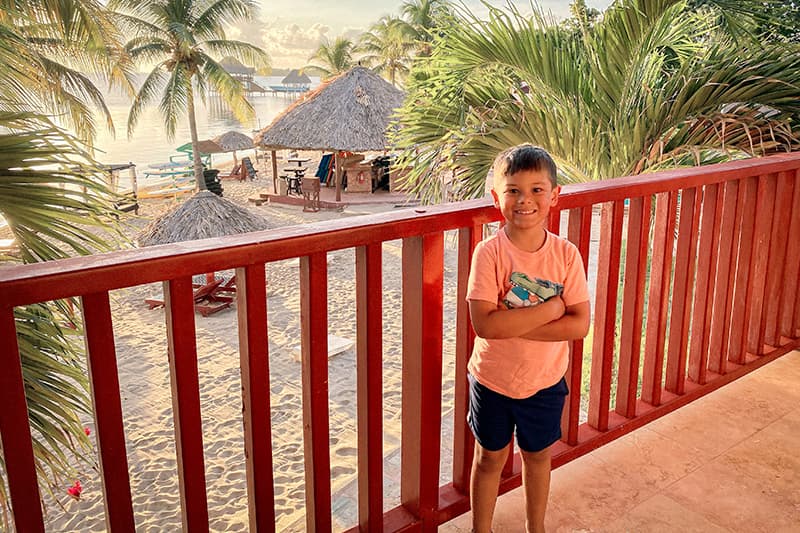 A smiling child stands on a balcony overlooking a sandy beach with palm trees and a thatched hut. A smiling child stands on a balcony overlooking a sandy beach with palm trees and a thatched hut.