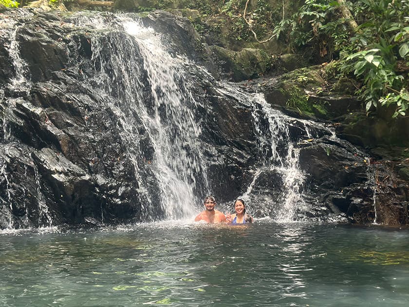 A couple stands in a natural pool beneath a cascading waterfall, surrounded by lush greenery. A couple stands in a natural pool beneath a cascading waterfall, surrounded by lush greenery.