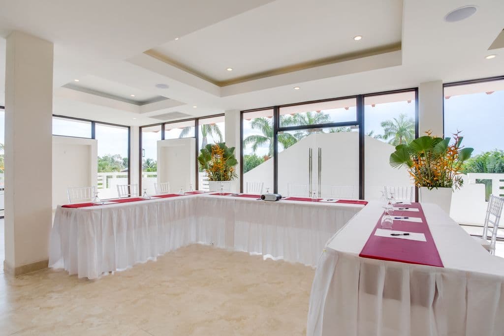 A spacious and modern meeting room with a U-shaped table arrangement. The room has a white tiled floor, white walls, and a high ceiling with exposed beams. There is a large palm tree in the corner, and a projection screen and projector at the front of the room. The table is covered in white tablecloths and has red runners. Clear plastic chairs are arranged around the table. There is a bar area with counter stools in the background.