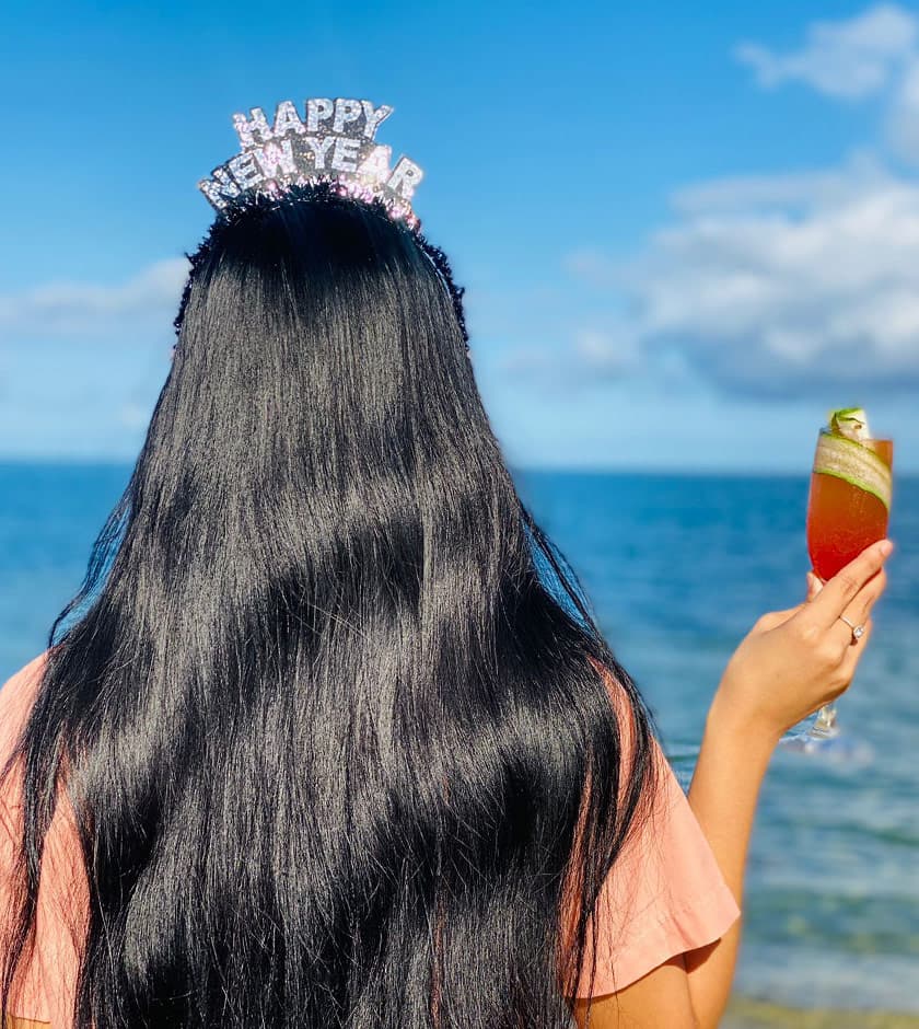 A person with long black hair holds a drink, wearing a "Happy New Year" headband by the ocean.