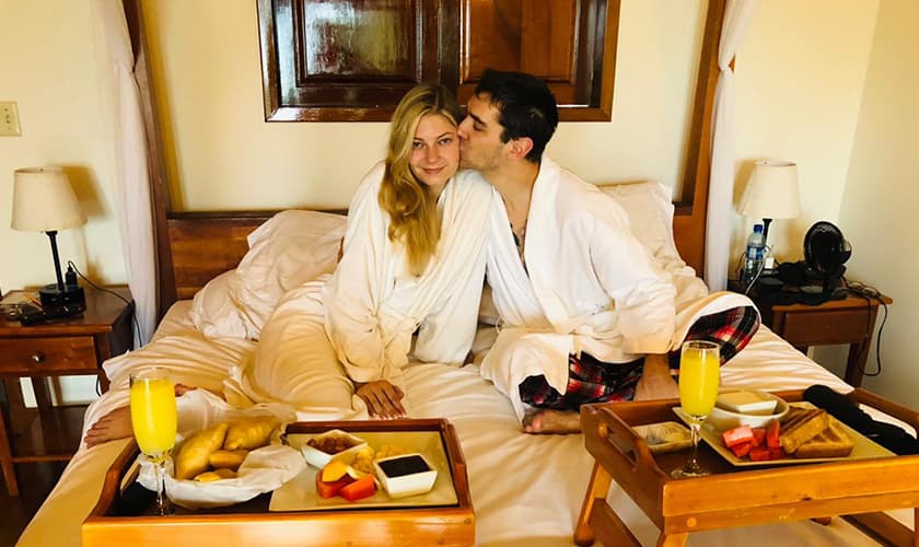 A couple in white robes sits on a bed with breakfast trays, sharing a kiss.