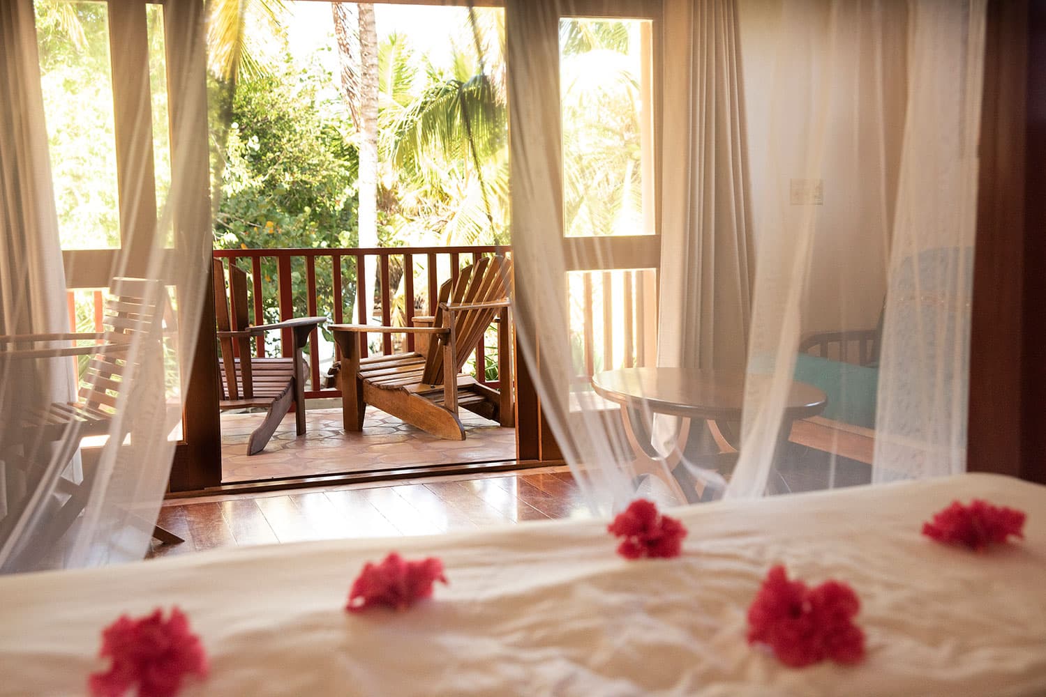 A bedroom with a king-sized bed and sheer white curtains. The bed is adorned with pink flower petals. Sliding glass doors lead to a private balcony with wooden chairs and a small table. Lush greenery and palm trees are visible outside.
