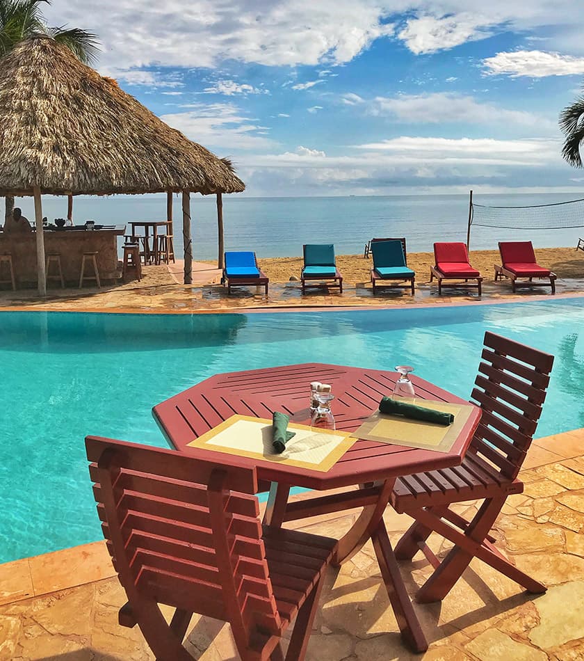 A poolside table overlooks a beach with colorful lounge chairs under a thatched roof.