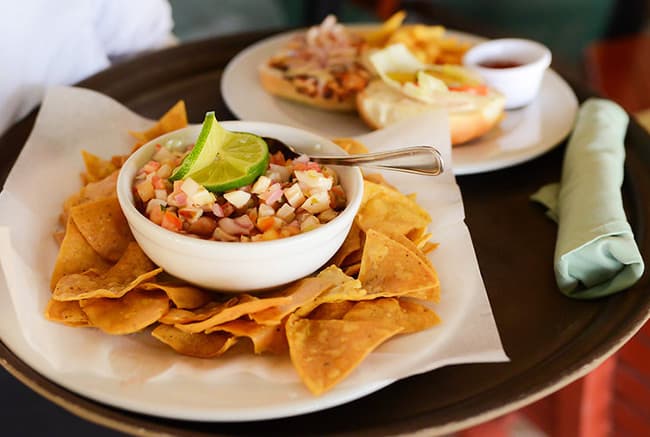 A tray of tortilla chips with a bowl of ceviche garnished with lime, alongside a plate of food in the background.