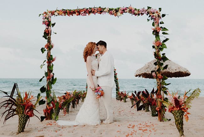 A bride and groom embrace under a floral arch on a beach.