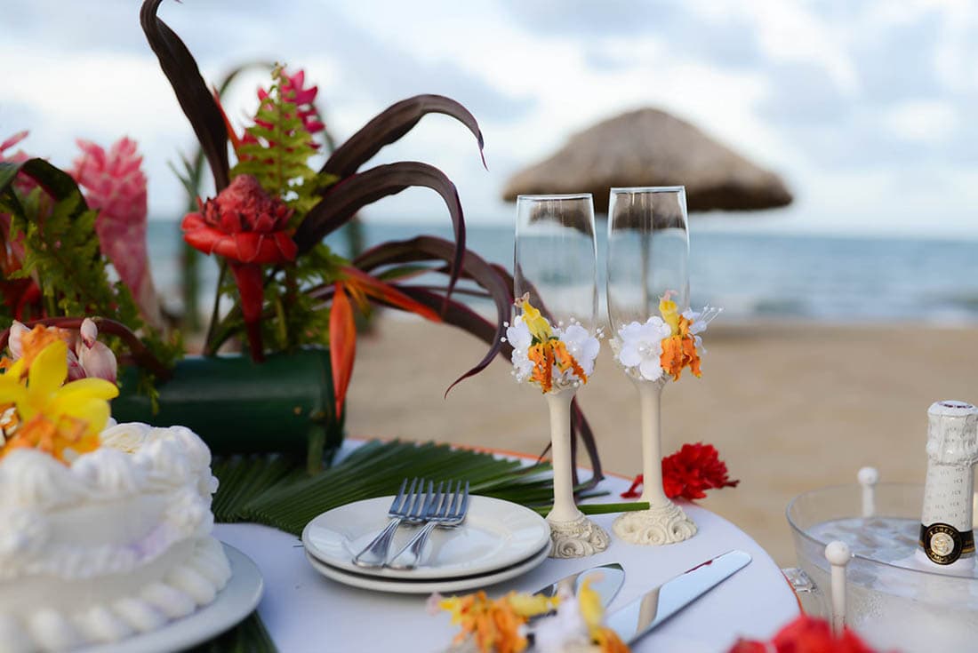 A collection of images showcasing a destination wedding in Belize. The images capture various moments of the wedding, including the ceremony, reception, and couple portraits. Highlights include a beach wedding with floral archways, a white wedding cake, and the couple walking hand-in-hand on the beach.