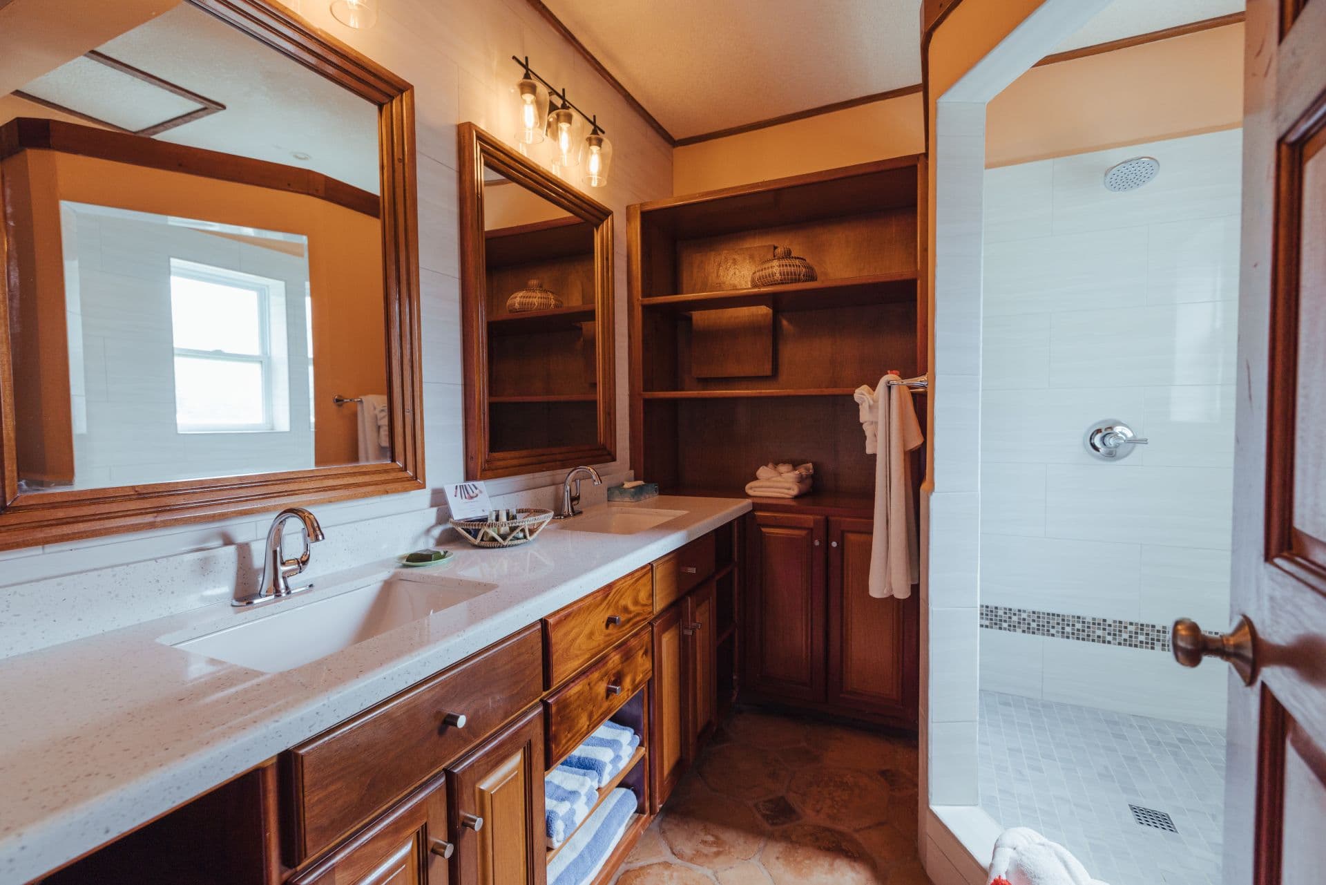 A spacious bathroom with dual sinks, a large mirror, and a walk-in shower. The walls are tiled with green and white tiles, and the floors are made of stone. There is a wooden vanity with drawers underneath the sinks, and a shelving unit with towels and other toiletries. The bathroom has a bright and airy feel with plenty of natural light. The sinks have colorful, patterned bowls.