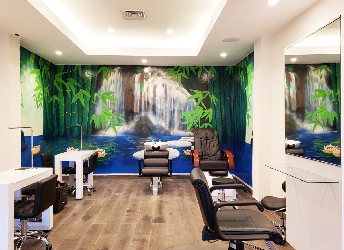 A modern nail salon with a calming mural of a waterfall, bamboo, and water lilies. There are multiple manicure stations with black chairs and white tables. A massage chair is visible in the background, and the overall atmosphere is serene and relaxing.