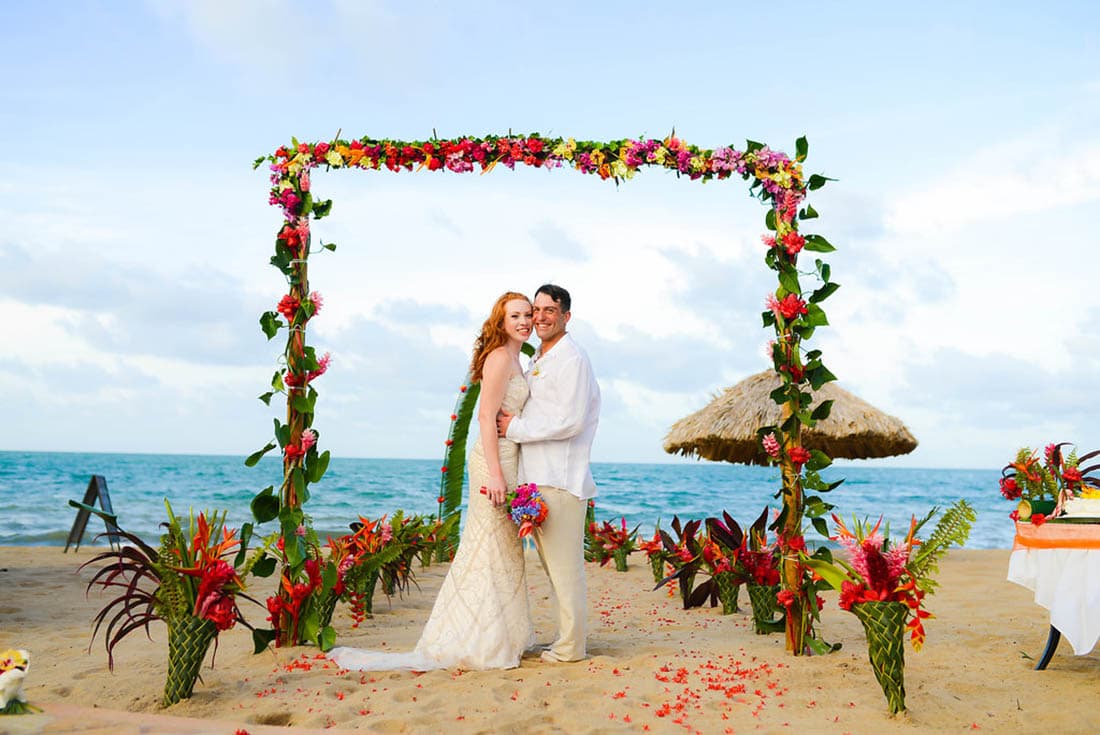 A red-haired bride and dark-haired groom pose under a floral wedding arch at the beach.