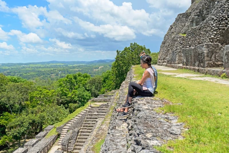 A person sits on a stone wall overlooking lush greenery and ancient structures under a bright blue sky.