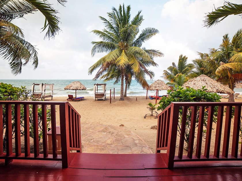 View of a sandy beach with palm trees and thatched cabanas, seen from a red wooden deck. View of a sandy beach with palm trees and thatched cabanas, seen from a red wooden deck.