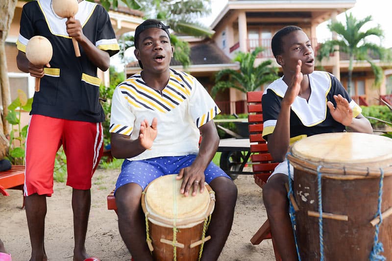 Three young men play drums and maracas while singing, surrounded by greenery and a beach setting.