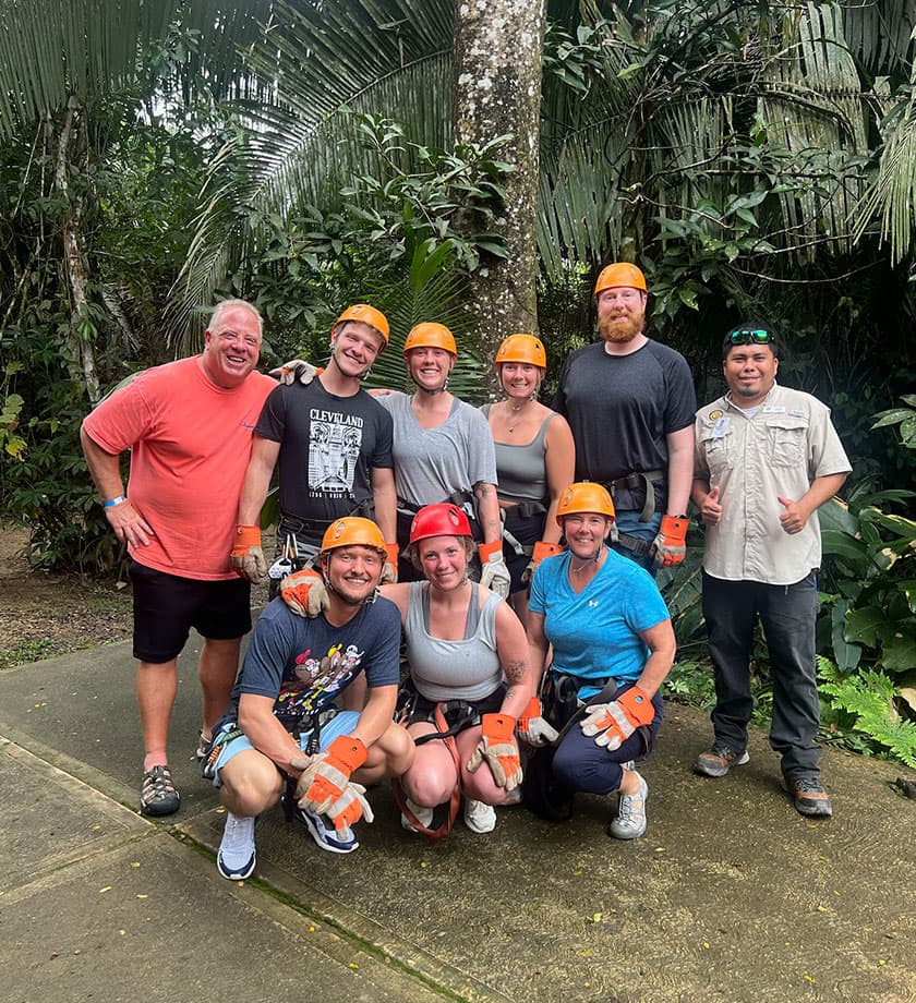 A group of smiling people wearing helmets and gloves poses together in a lush, tropical setting.