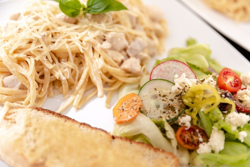 A plate of fettuccine Alfredo with chicken, served with a colorful side salad and garlic bread.