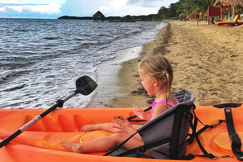 A young girl sits in an orange kayak on a sandy beach, gazing at the water.