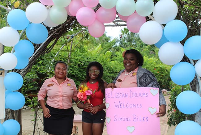 Three women, including one holding flowers, stand under a festive arch with balloons, welcoming Simone Biles with a sign.