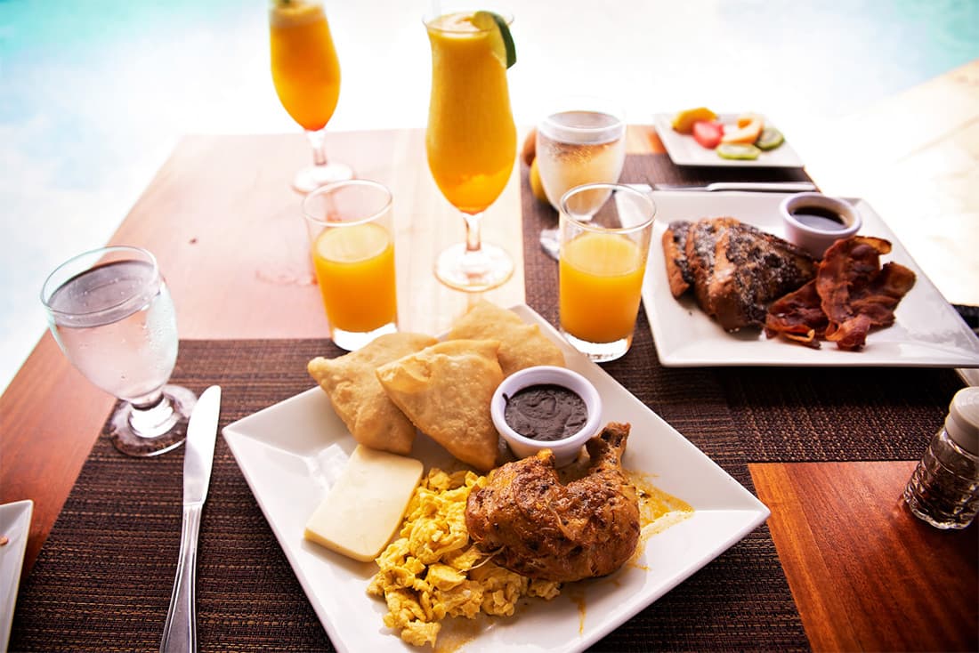 A delicious breakfast spread on a table overlooking a pool. The meal includes scrambled eggs, fried chicken, johnnycakes, cheese, bacon, fruit, and several glasses of orange juice and water.