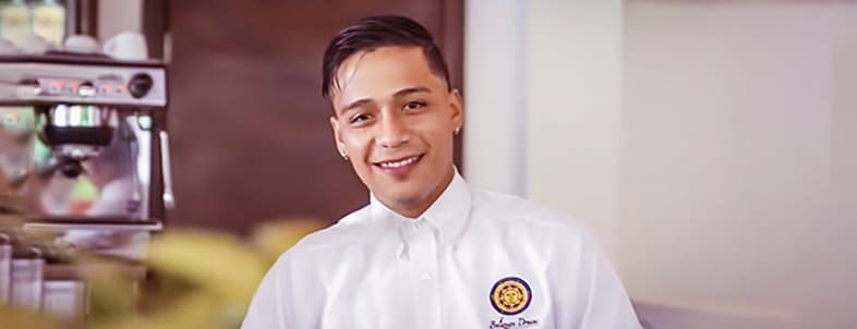 A smiling barista in a white shirt stands behind a coffee counter. A smiling barista in a white shirt stands behind a coffee counter.