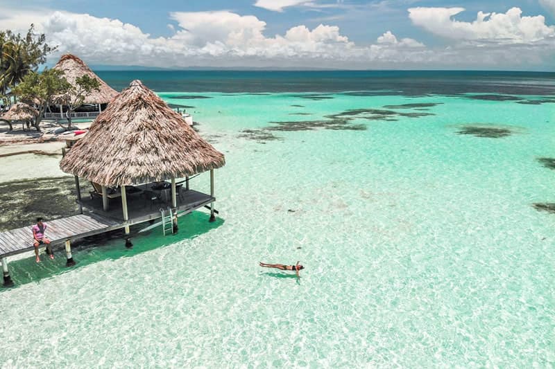Aerial view of a crystal-clear lagoon with a thatched-roof hut and a person swimming.