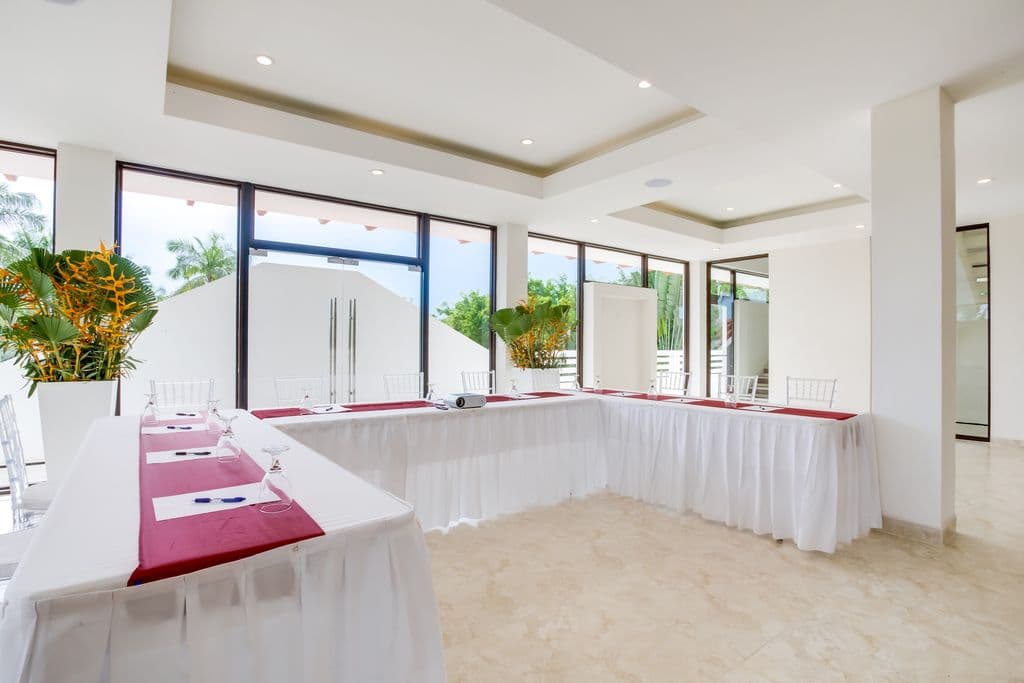 A modern meeting room with a U-shaped table arrangement. The room has a white tiled floor, white walls, and a high ceiling with exposed beams. A large projection screen and projector are at the front of the room. The table is covered in white tablecloths with red runners. Clear plastic chairs are arranged around the table. There is a bar area with counter stools in the background.