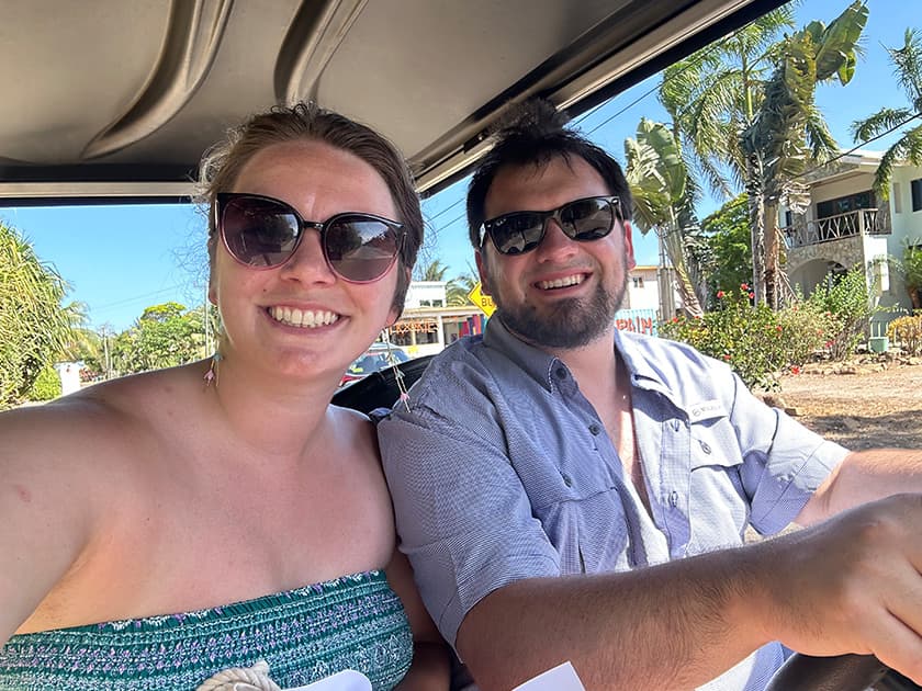 A smiling couple wearing sunglasses poses for a selfie in a golf cart on a sunny day. A smiling couple wearing sunglasses poses for a selfie in a golf cart on a sunny day.