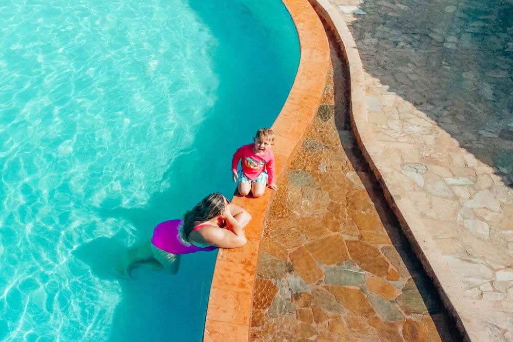 A woman in a pink bathing suit leans on the edge of the sparkling pool talking to a happy young boy in a rash guard shirt kneeling on the pool surround.