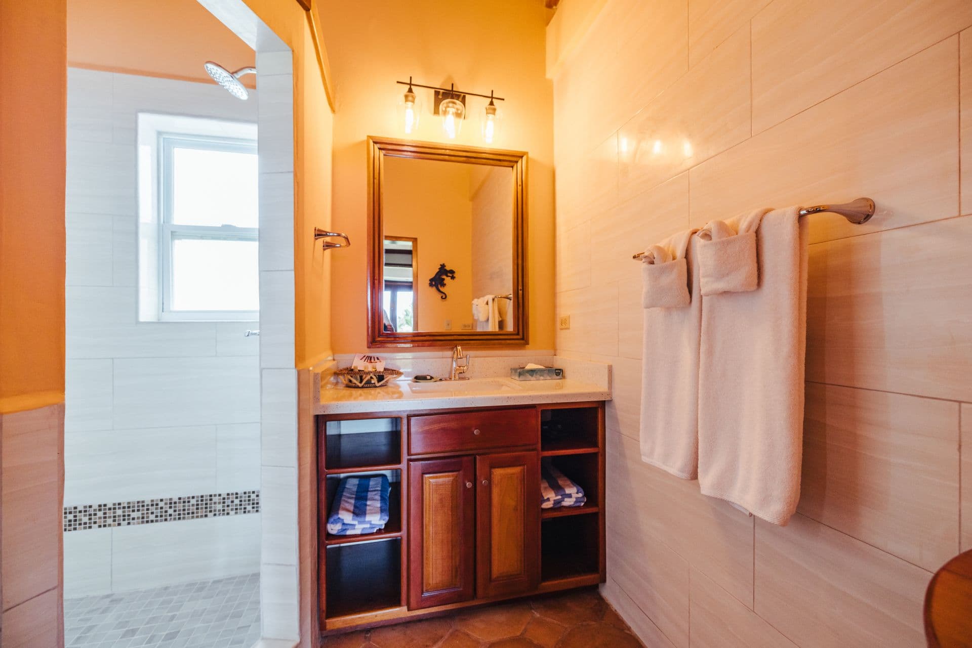 A spacious bathroom with dual sinks, a large mirror, and a walk-in shower. The walls are tiled, and the floors are made of stone. There is a wooden vanity with drawers underneath the sinks, and a shelving unit with towels and other toiletries. The bathroom has a bright and airy feel with plenty of natural light.