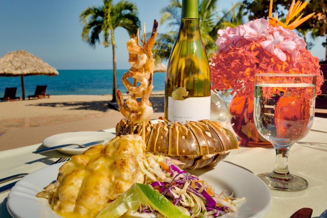 A plate of fish and chips served on a bed of parchment paper. The fish is breaded and fried, and it's accompanied by a small bowl of tartar sauce, a wedge of lime, and a pile of golden French fries. A glass of lemonade and a potted plant are visible in the background.