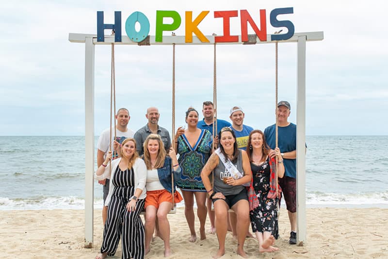 A group of ten people poses together on a beach under a colorful sign that reads "HOPKINS."