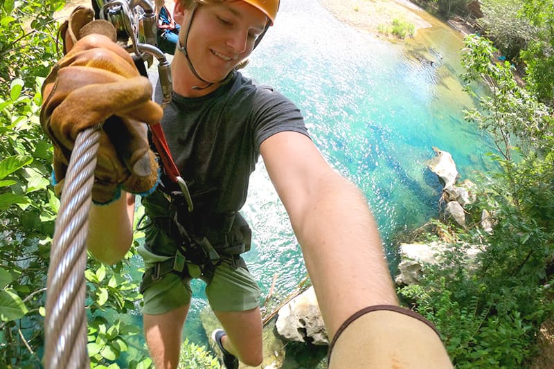 A person in safety gear is rappelling down near a blue river surrounded by lush greenery. A person in safety gear is rappelling down near a blue river surrounded by lush greenery.