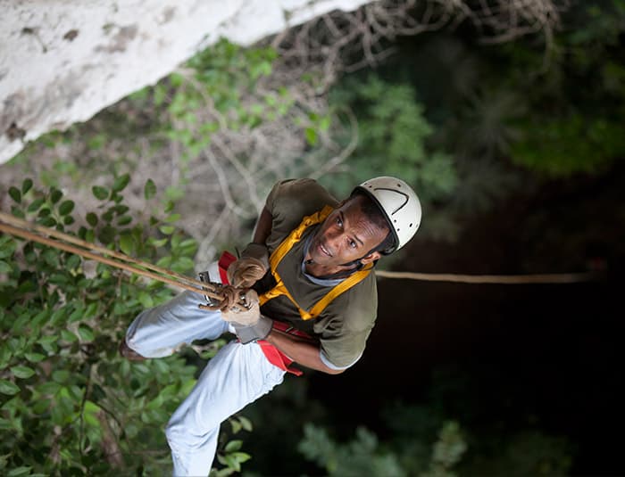 A man in a brown tee, white helmet and pants descends on a rappelling line into a deep ground cave.